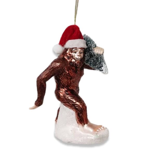 Glass Christmas ornament of Bigfoot wearing a Santa hat and carrying a christmas tree